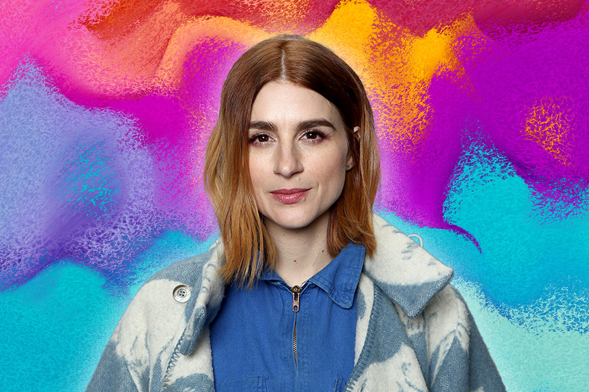 Aya Cash wears a blue and white-washed denim jacket over a blue zipped up shirt. She stares at camera. Behind her is a blue, purple, yellow and red ombre background.