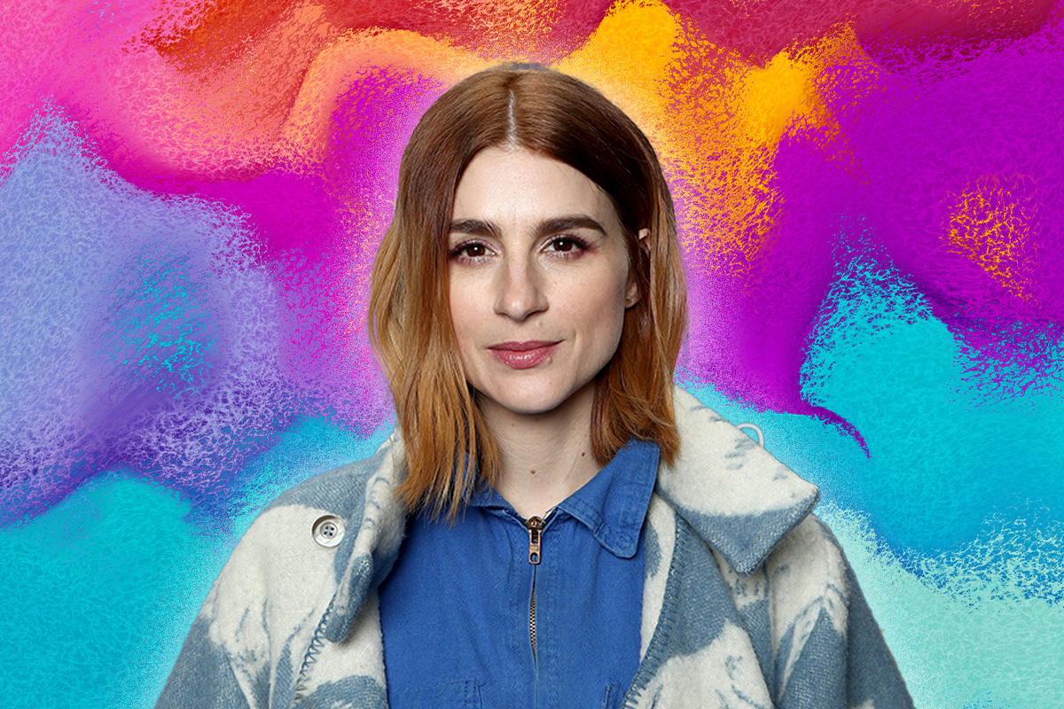 Aya Cash wears a blue and white-washed denim jacket over a blue zipped up shirt. She stares at camera. Behind her is a blue, purple, yellow and red ombre background.