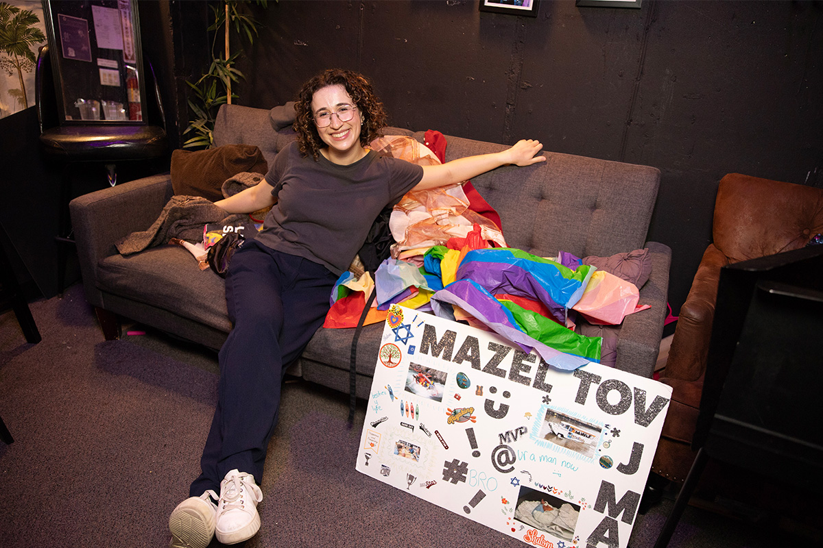 Lily Blumkin smiles at camera. She sits on a couch with colorful props. There's a poster board reading, "Mazel Tov, J-Man" is at her feet.