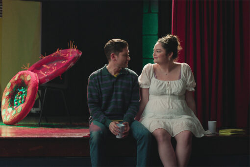 A man and a woman sit on the edge of a stage looking at each other.