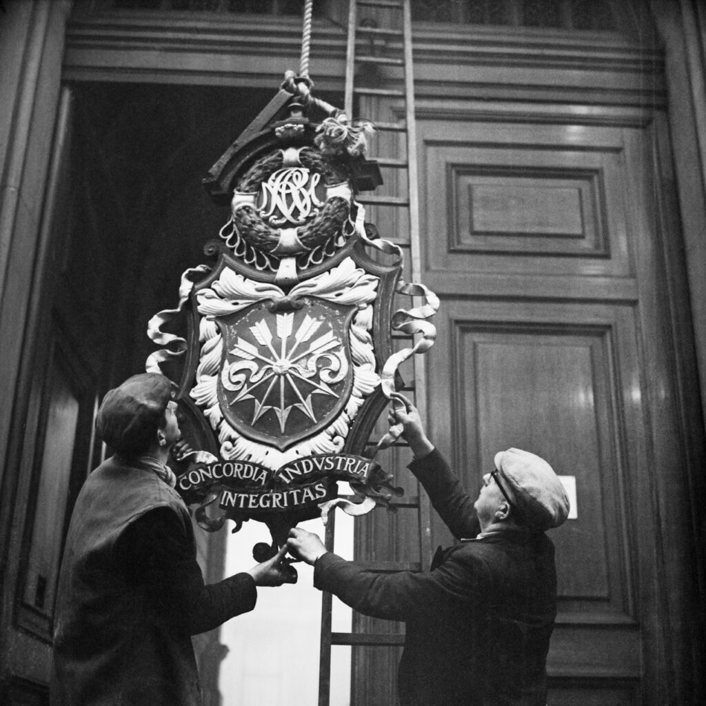 Two men support the insignia of NM Rothschild & Sons at New Court, as it is lowered in the headquarters of the Rothschild investment bank, on St Swithin's Lane in the City of London, England, 5th December 1962. The insignia incorporates the Rothschild's 'Five Arrows' (each arrow represents a son of Mayer Amschel Rothschild and their respective businesses).