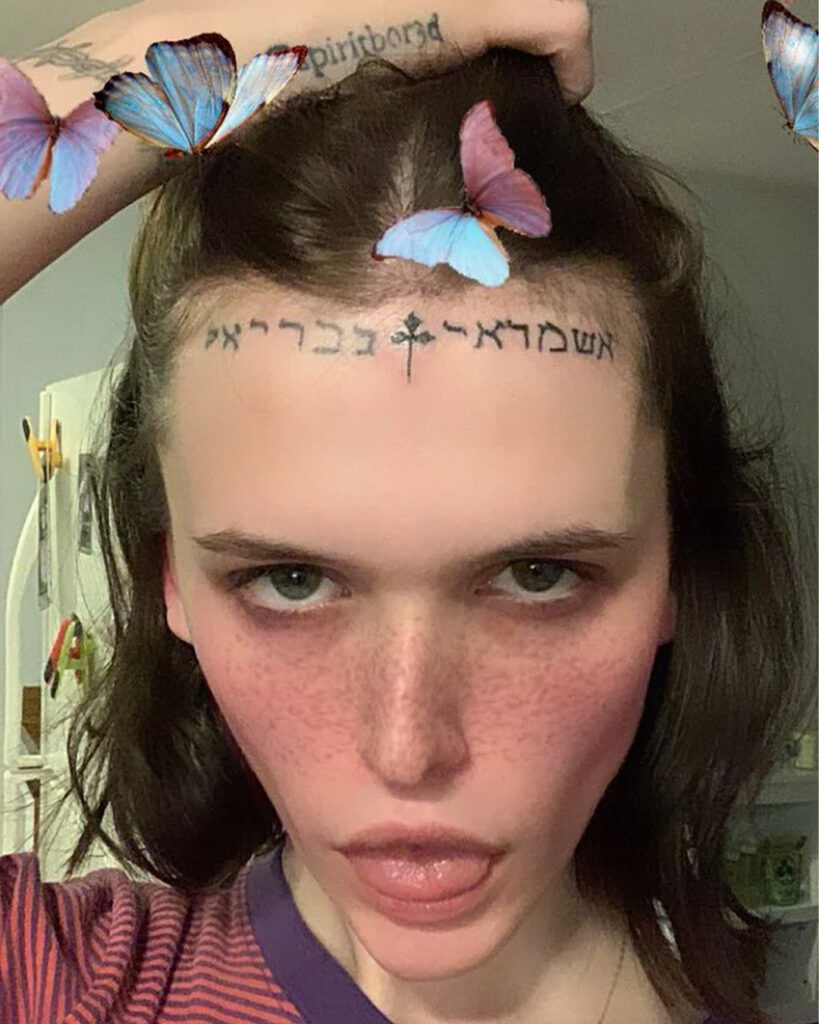 A woman stares at camera. She has a tattoo in Hebrew lettering on her forehead.
