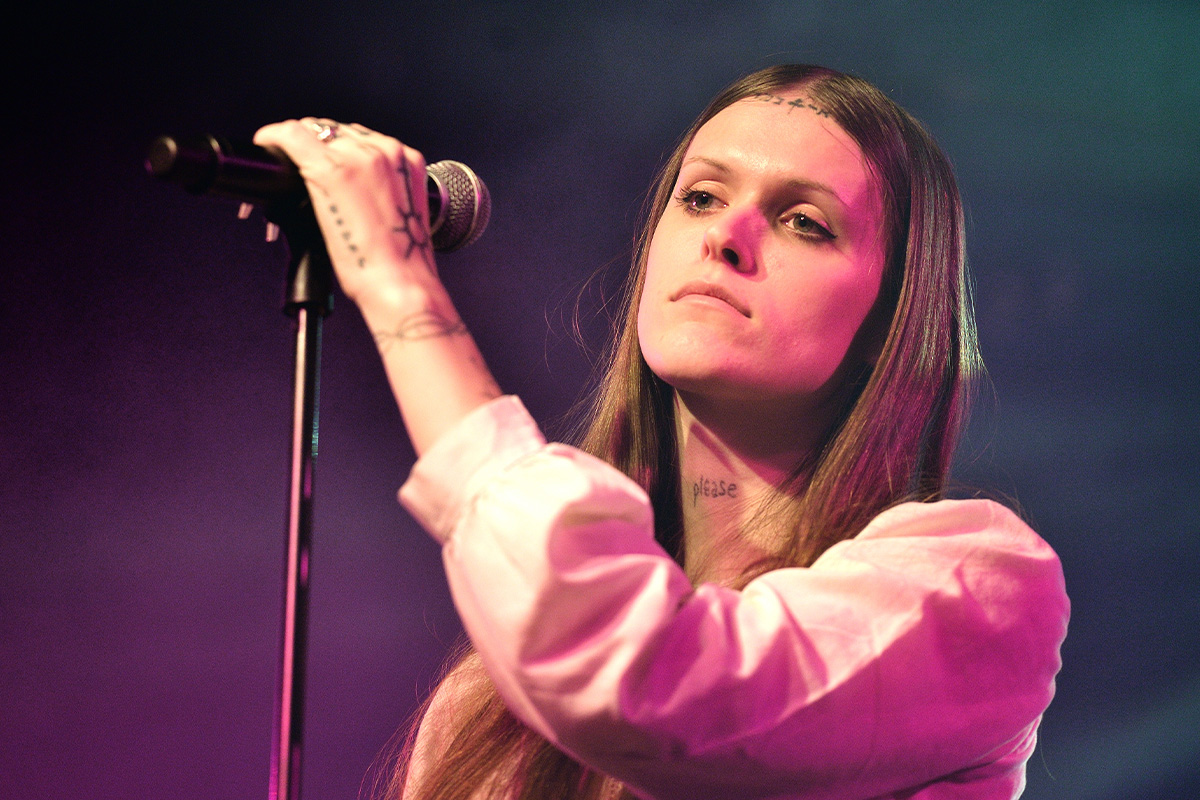A woman, Ethel Cain, with tattoos on her hands and at her hairline holds a microphone and stares off into the distance.