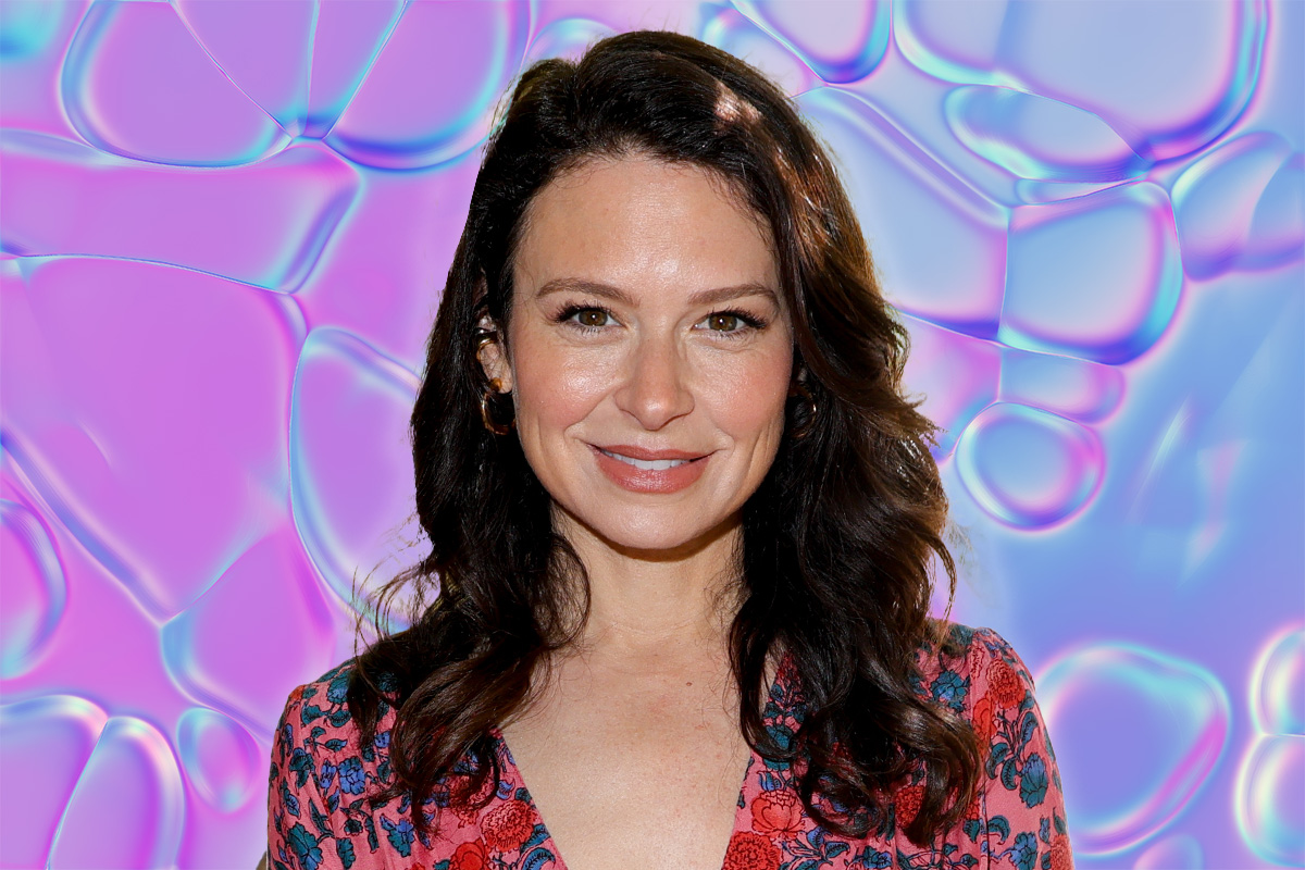 Katie Lowes smiles at camera with a colorful pink, blue and purple background behind her.