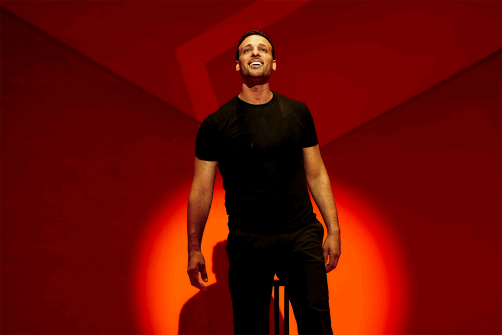 Ari'el Stachel in "Other" Off-Broadway. He smiles and looks up as a spotlight shines on him. The set behind him is reddish orange.