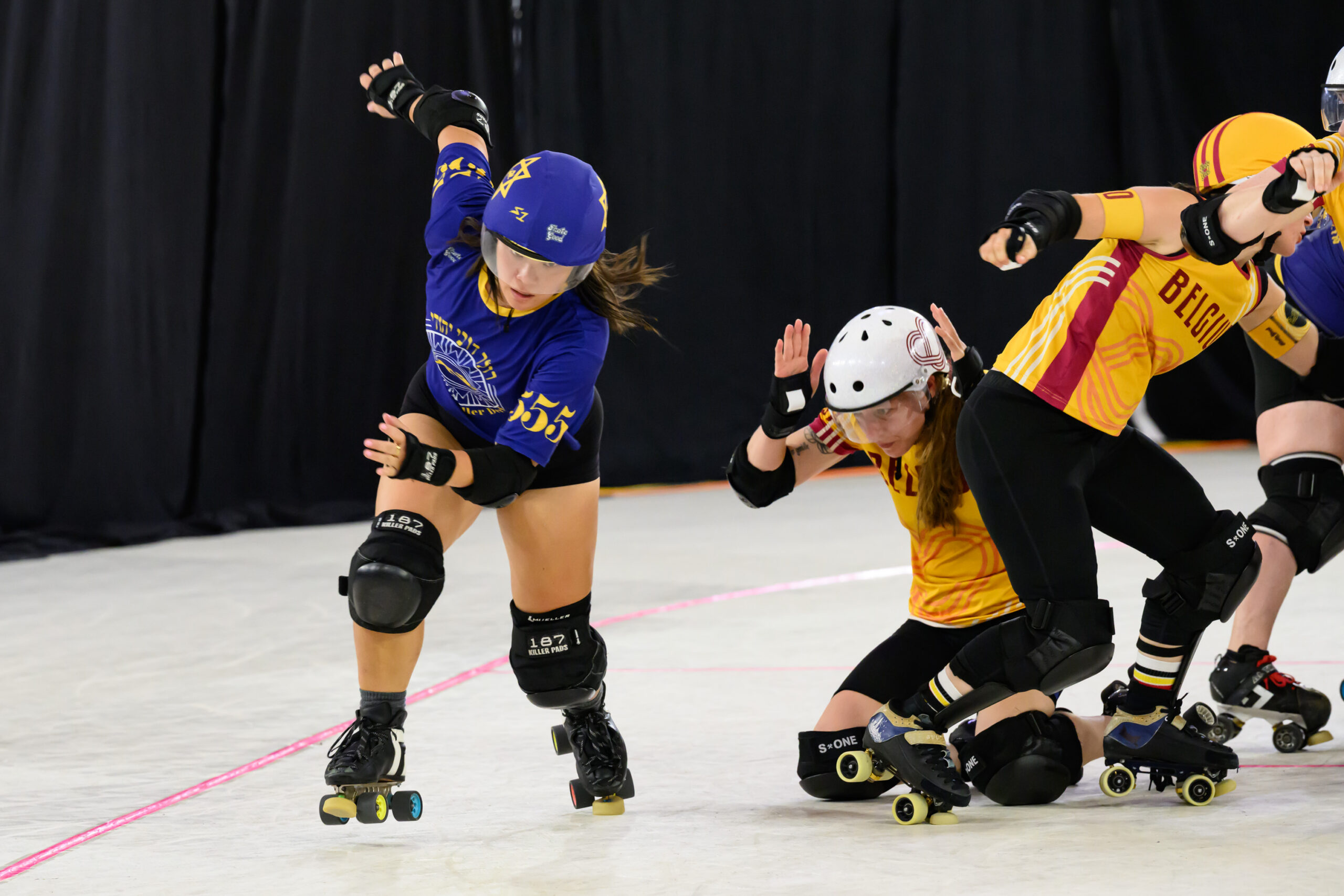 A Jewish roller derby skater in blue skates past a player from Team Belgium, who is on their knees with their hands up at the 2025 Roller Derby World Cup.